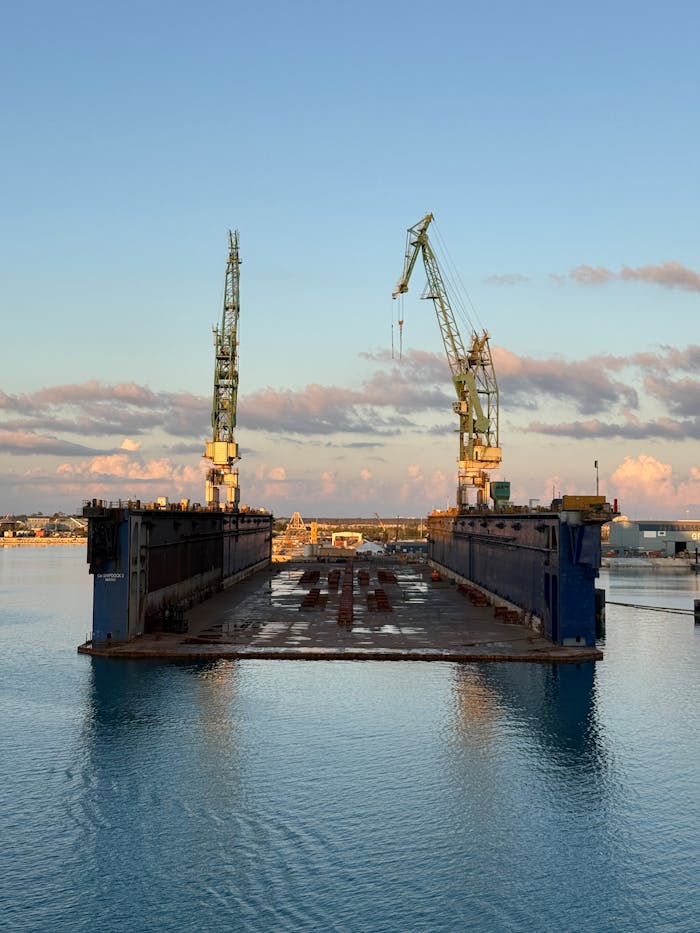 A floating dock flanked by cranes in a calm port at sunrise, with clouds in the sky.